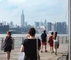 People are walking along a riverside boardwalk with a view of the New York City skyline in the background