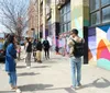 A group of people is attentively listening to a person facing them possibly a guide on a sunny day in front of a building with colorful street murals