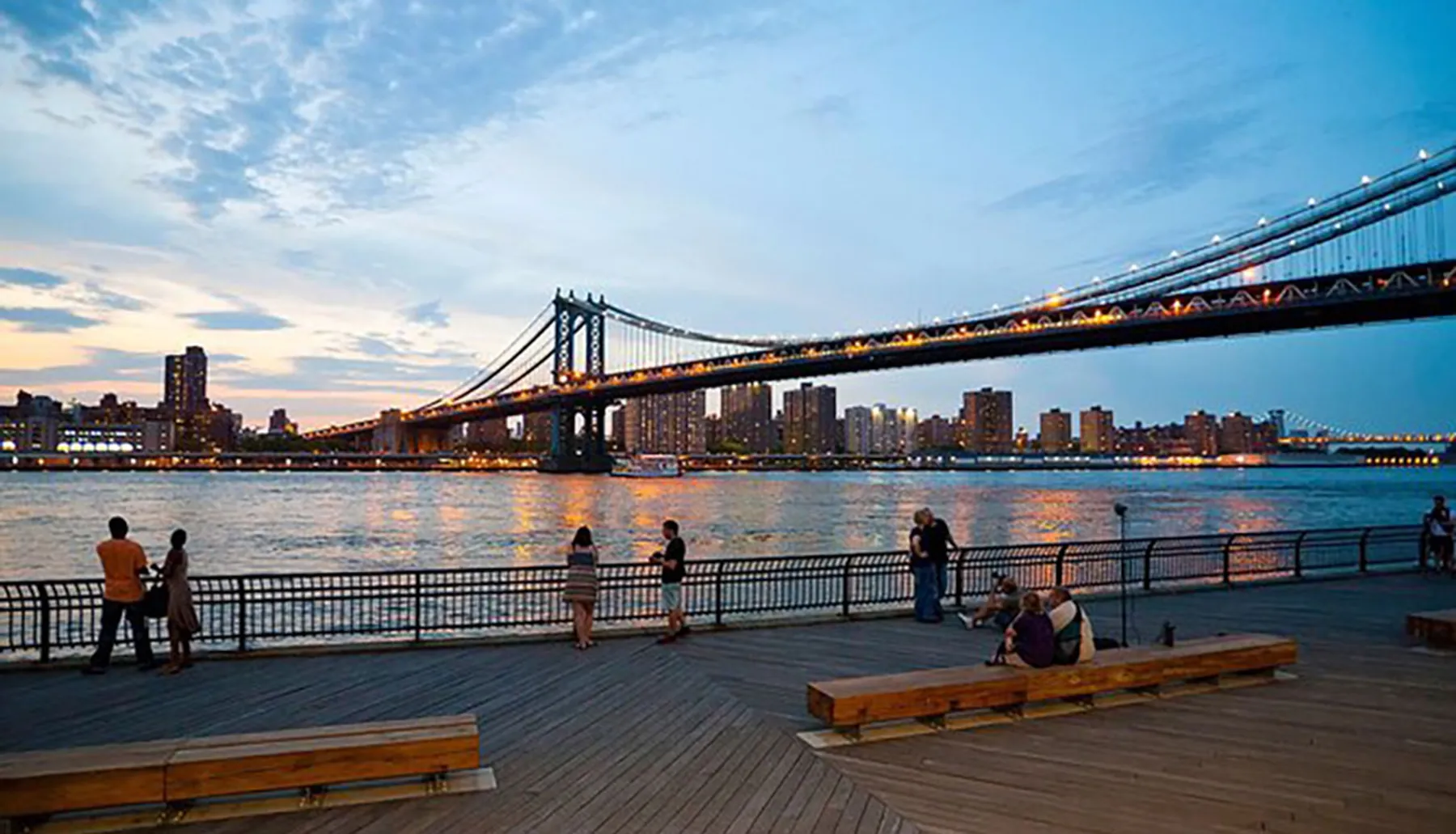 People enjoy the evening view of a lit-up suspension bridge from a waterfront boardwalk.