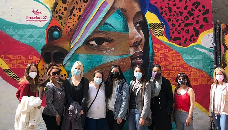 A group of people wearing masks pose in front of a colorful mural featuring a large portrait of a woman's face.