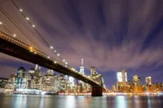 The image shows a long exposure night shot of the Brooklyn Bridge with radiant stars of light and streaky clouds above, set against the backdrop of the illuminated New York City skyline.