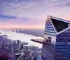 The image shows a modern skyscraper with a distinctive angled top overlooking a panoramic view of a citys skyline during what appears to be dawn or dusk with a river and other tall buildings in the background