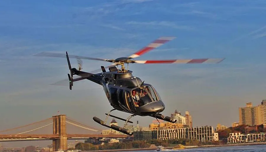 A helicopter is flying over a city skyline near a large bridge during what appears to be clear weather conditions.