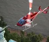 A red helicopter is flying near the torch of the Statue of Liberty against a background of water