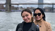 Two smiling individuals pose for a photo with a prominent bridge in the background, likely taken on a boat or waterfront.