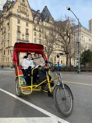 Two individuals are smiling for a photo while seated in a red and yellow pedicab in front of a historic building with leafless trees and a streetlamp in the background.