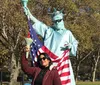 A person is posing with a person dressed as the Statue of Liberty who is holding an American flag in a park setting with trees in the background