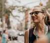 The image shows a smiling woman with glasses looking to her right on a busy city street with blurred pedestrians and traffic in the background