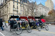 A group of people enjoys a pedal-powered rickshaw ride in an urban park setting with historic buildings in the background.