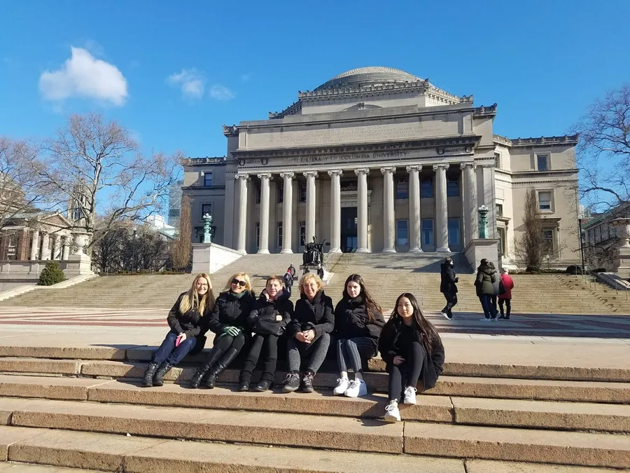 A group of people is sitting on the steps in front of the neoclassical facade of the Low Memorial Library at Columbia University on a sunny day.
