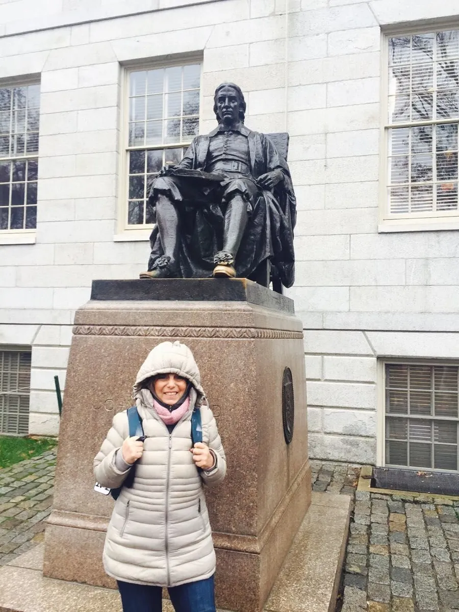 A person wearing a hooded coat and a backpack is smiling with thumbs up in front of a seated bronze statue of a historical figure on a stone pedestal.