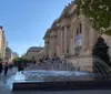 The image shows a bustling scene outside the Metropolitan Museum of Art with visitors and a fountain in the foreground on a bright sunny day