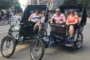Three people are enjoying a ride through an urban street in pedal-powered rickshaws.