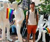 A smiling person stands beside white statues of people one of which is holding a rainbow pride flag in what appears to be a vibrant outdoor setting