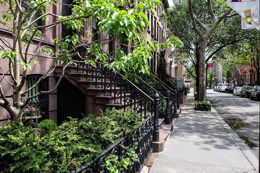 The image shows a tree-lined urban street with classic brownstone townhouses, featuring stoops with wrought-iron railings, lush greenery, and a clear blue sky peeking through the foliage.