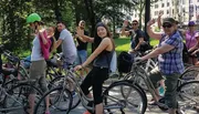 A group of cheerful people on bicycles are posing for a photo, waving and smiling in a park-like setting.
