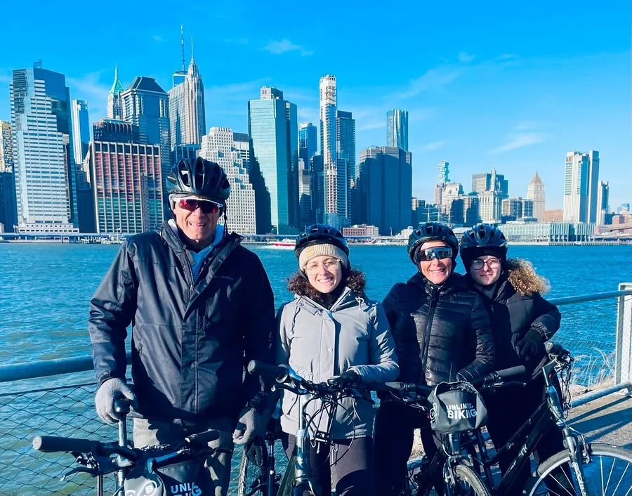 Four cyclists wearing helmets are posing for a photo with a city skyline in the background.