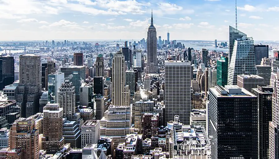 An aerial view of the Manhattan skyline with the Empire State Building in the center, set against a backdrop of blue skies and scattered clouds.