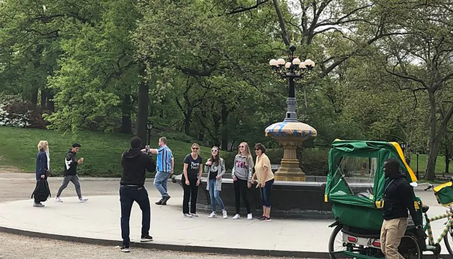 A group of people are posing for a photo taken by a man in a black hoodie in what appears to be a park, with a lamppost in the background and a pedicab on the right.