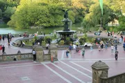 The image shows a lively scene of people gathering and walking around the Bethesda Terrace and Fountain in Central Park, New York City, on a sunny day.