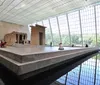 The image shows an ancient Egyptian temple structure exhibited within a large light-filled room featuring a glass wall and a reflecting pool with visitors observing and resting around the space