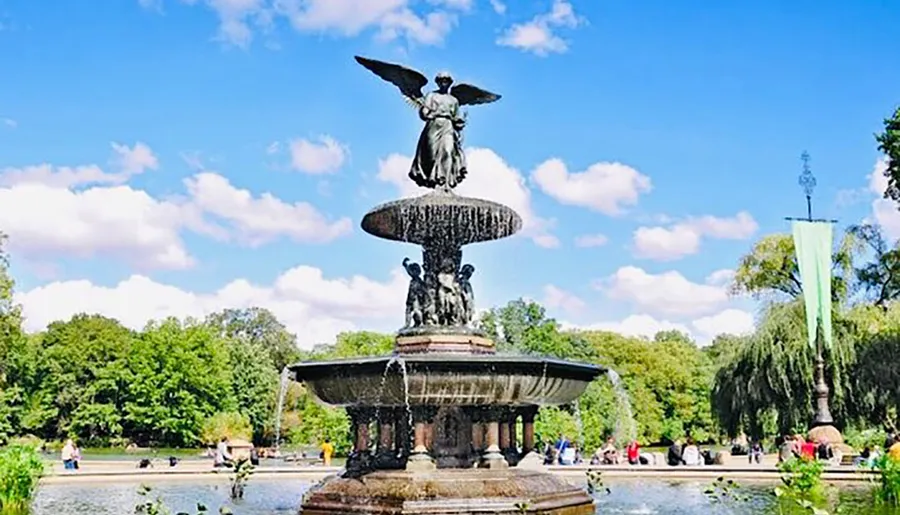 The image shows a grand fountain with water cascading from multiple tiers and a statue at the top, set in a sunny park with people leisurely gathered around.