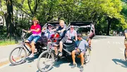 A group of people is posing for a photo with smiles on a pedal-powered vehicle, likely a pedicab, on a sunny day with trees in the background.