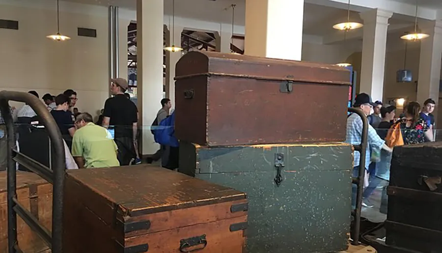The image shows a collection of vintage trunks displayed in a room with people milling about in the background, suggesting a museum or exhibition setting.