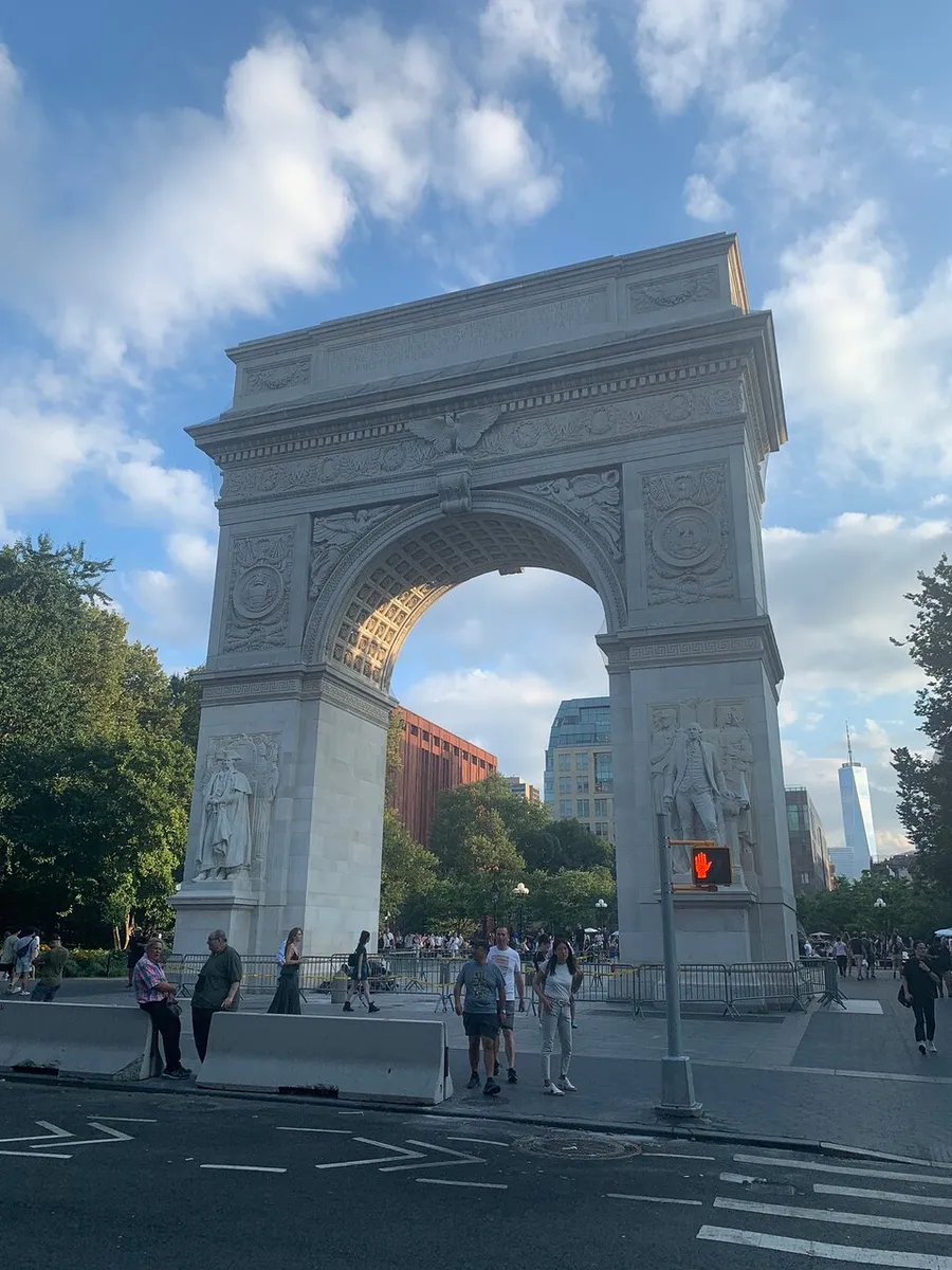 The image shows the Washington Square Arch in New York with people walking around and the One World Trade Center visible in the background under a partly cloudy sky.