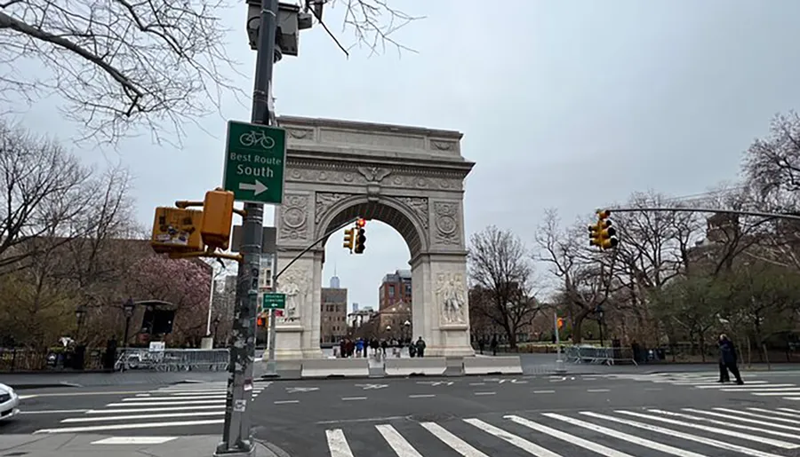 The image shows the Washington Square Arch located at the northern entrance of Washington Square Park in Manhattan, with a bicycle route sign in the foreground and a skyscraper faintly visible in the distance.