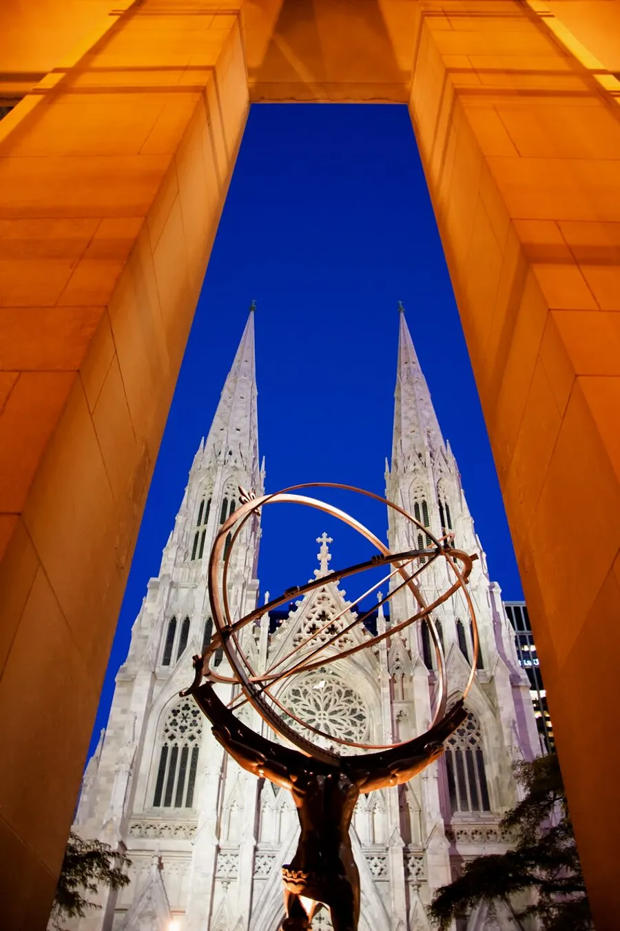 The image shows the towering spires of a Gothic cathedral framed by a dramatic archway and contrasted with a modern sculpture in the foreground against a deep blue sky.