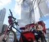 A child and an adult sit in a red bicycle trailer with a bicycle in the foreground and skyscrapers in the background under a blue sky