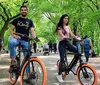Two people are smiling and riding electric bikes in a tree-lined park with other park-goers in the background
