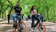 Two people are smiling and riding electric bikes in a tree-lined park with other park-goers in the background.