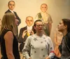 Three women are engaged in a conversation in an art gallery with surreal paintings of figures with oversized heads in the background