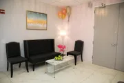 A modern waiting room with a black couch and chairs, a white marble table with fruit on it, pink flowers, balloons, and a painting on the wall.