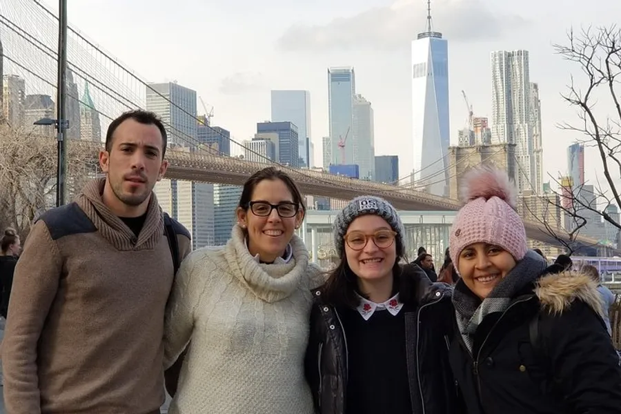 Four people are posing for a photo with the Brooklyn Bridge and the New York City skyline, including the One World Trade Center, in the background.