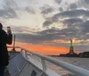 Two people are taking photos with their smartphones of the Statue of Liberty against a beautiful sunset sky from a boat