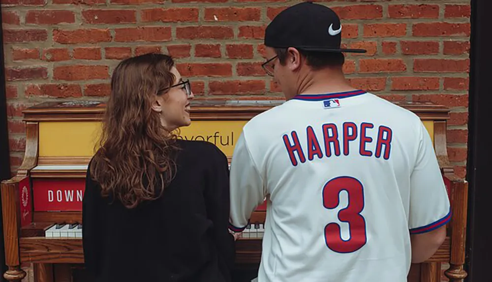 Two people are facing each other smiling beside a piano against a brick wall background with one wearing a baseball jersey with HARPER 3 on the back