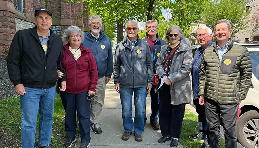 A group of eight smiling people, some wearing badges and casual attire, stand together on a sidewalk in a leafy suburban area.