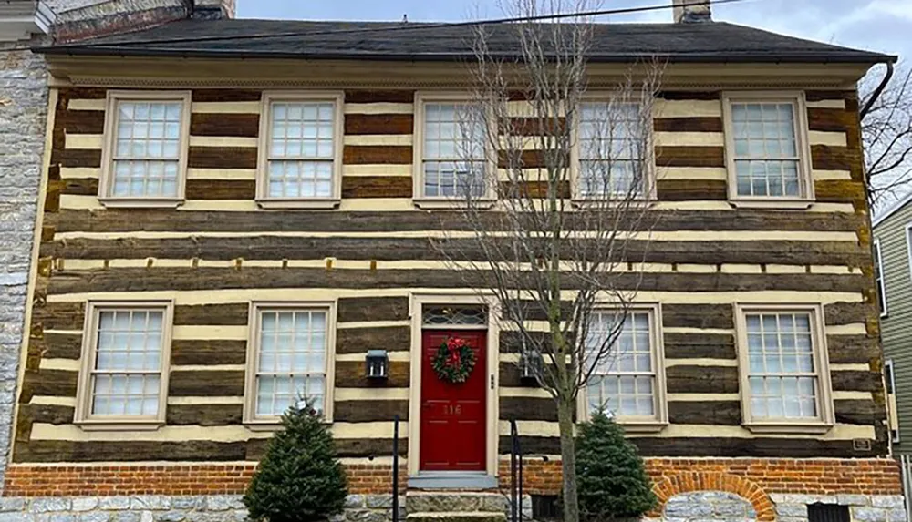 The image shows a two-story log cabin with a red front door decorated with a Christmas wreath and has a brick foundation