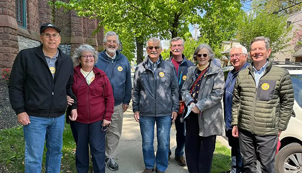 A group of smiling adults wearing matching circular stickers stands on a sidewalk with trees and a brick building in the background