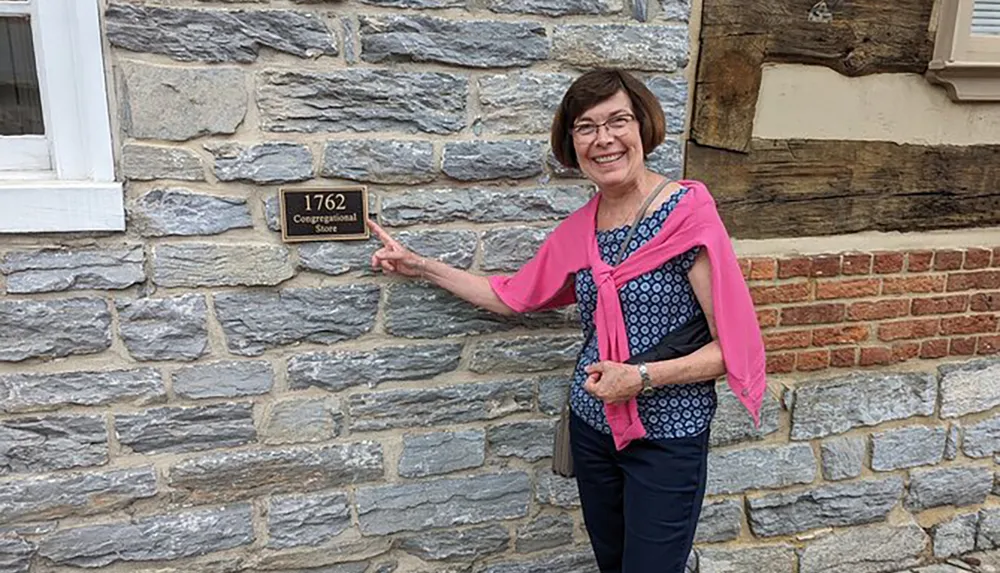 A smiling woman is pointing at a historical plaque on a stone building that reads 1762 Congressional Store