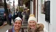 Two smiling women pose for a photo on a busy street with a historical plaque on the wall, outdoor seating, pedestrians, and an Amazon delivery van in the background.