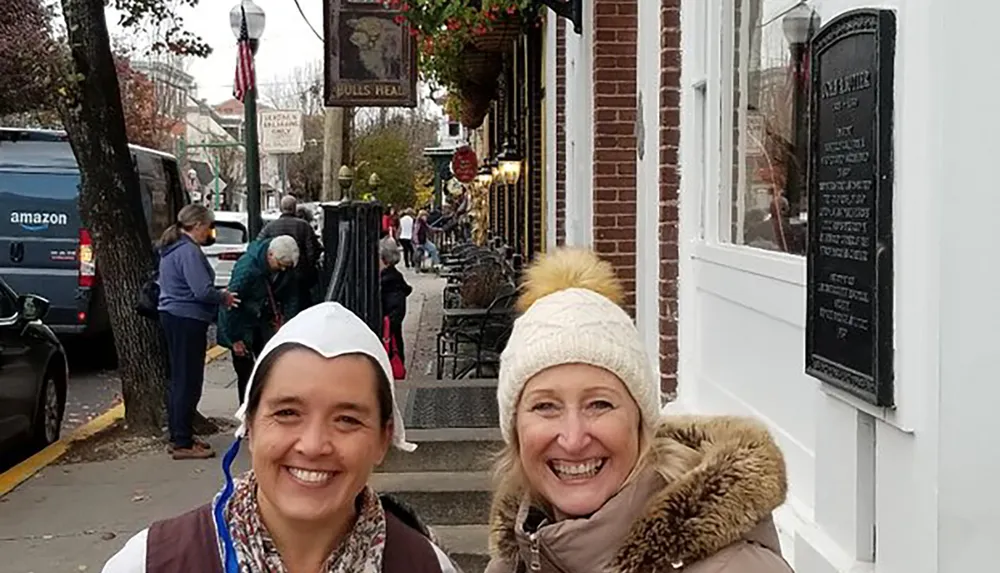 Two smiling women pose for a photo on a busy street with a historical plaque on the wall outdoor seating pedestrians and an Amazon delivery van in the background