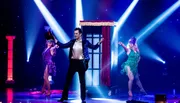 Two performers in sparkly, cabaret-style costumes strike poses on a stage bathed in blue and white spotlight beams.