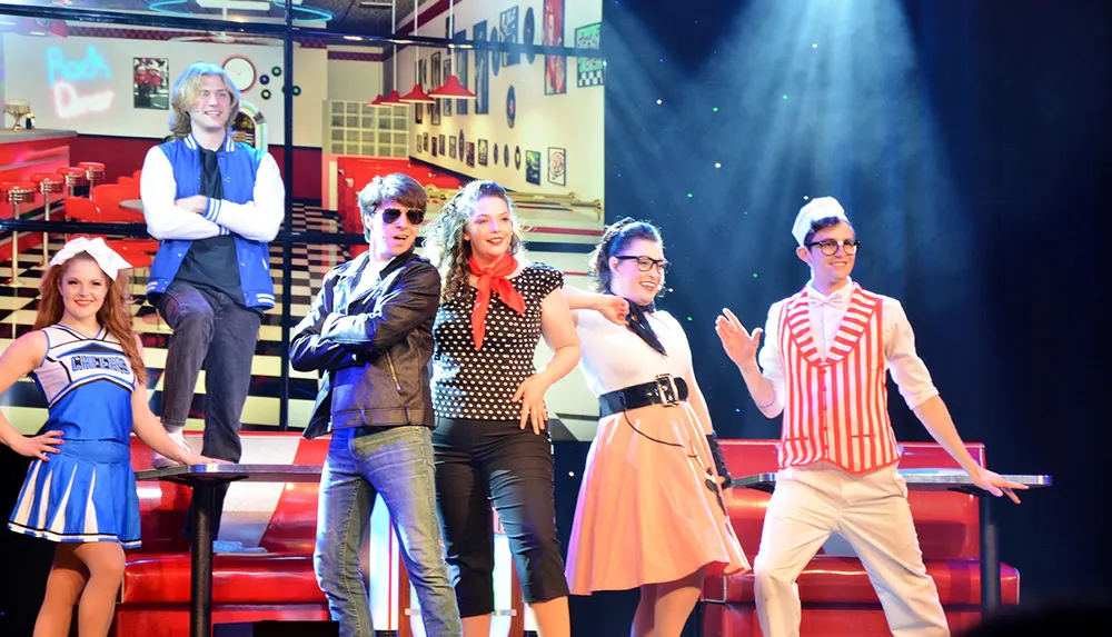 A group of people are dressed in 1950s American high school-themed costumes posing on a stage set that resembles a diner with a Rock Diner sign in the background suggesting a musical or theatrical performance
