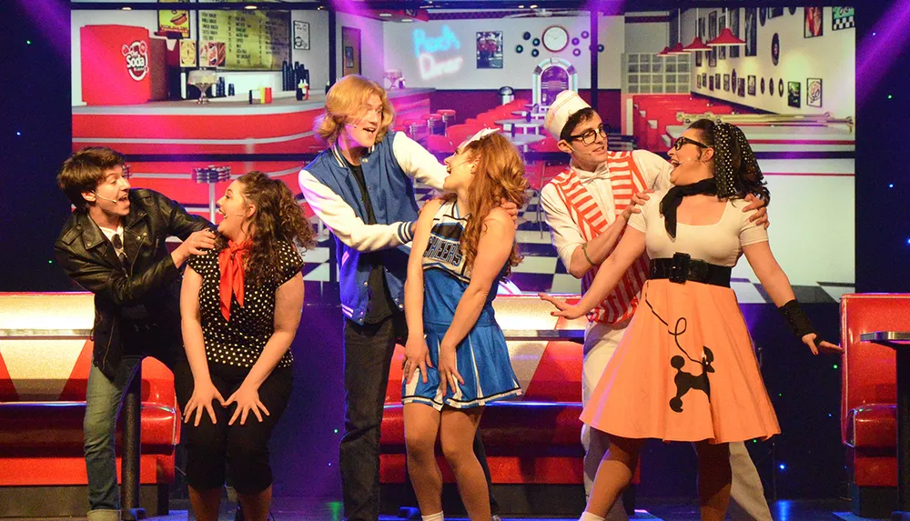 The image depicts a lively group of performers dressed in 1950s style attire acting out a scene on a retro-themed stage set to resemble a diner