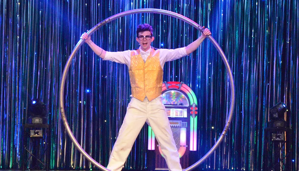 A person in a white outfit and glasses striking a pose with a large hoop on stage with a shimmering blue background and a jukebox to the side