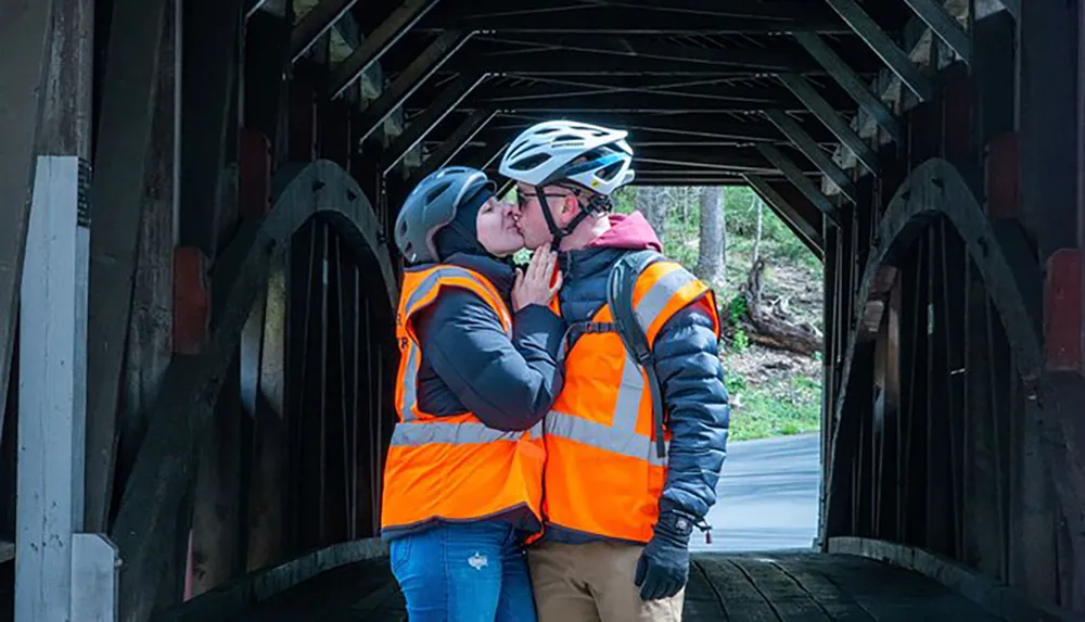 Two people wearing reflective vests and bike helmets are kissing inside a covered bridge